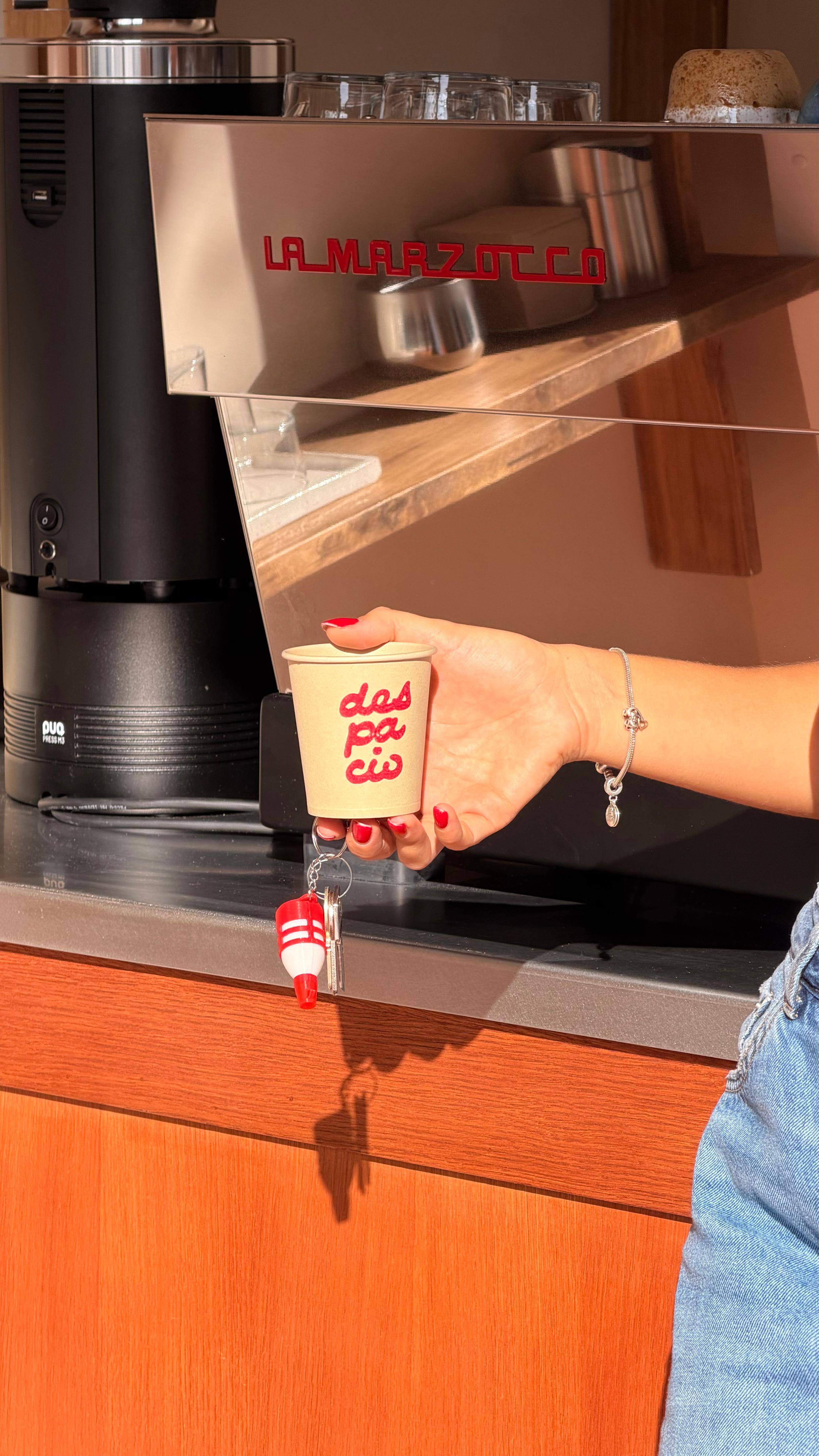 Una mano sostiene un vaso de Despacio frente a la máquina de espresso